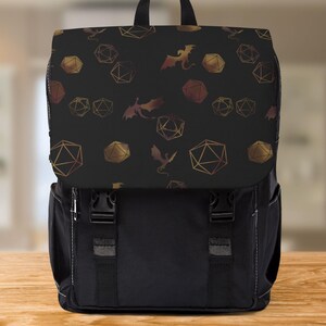 Può includere: Zaino nero con patta con un motivo di dadi geometrici e draghi dorati e bronzo. Lo zaino ha un corpo nero con due cinghie anteriori e una maniglia superiore. La borsa è su una superficie di legno.