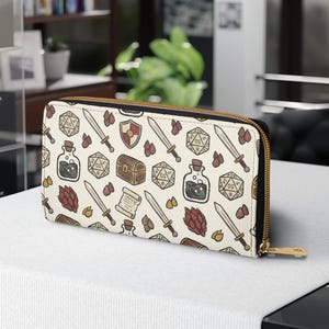 May include: A rectangular zip-around wallet featuring an off-white background with a repeating pattern of fantasy-themed illustrations including swords, shields, treasure chests, scrolls, potion bottles, and multi-sided gaming dice in muted colours.