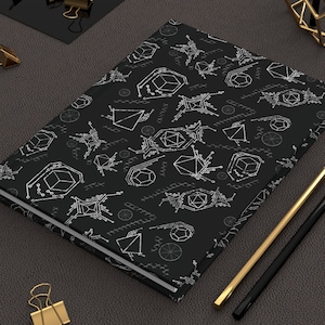 May include: A black notebook with a white geometric pattern of various shapes, including circles, triangles, and diamonds. The notebook is lying on a dark brown surface with two pencils and a few gold paper clips.