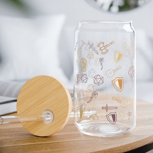 Könnte beinhalten: Eine durchsichtige Glasdose mit Bambusdeckel und Strohhalm. Die Dose ist mit einem Muster aus Würfeln, Schwertern, Schilden und anderen Fantasy-Symbolen verziert.