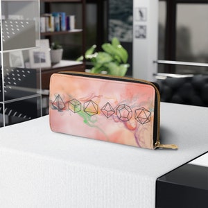 Könnte beinhalten: Eine lange Geldbörse mit Reißverschluss und farbenfrohem, abstraktem Design. Die Geldbörse zeigt eine Reihe schwarzer geometrischer Formen, die Würfeln ähneln, vor einem Hintergrund aus rosa, orange und grünen Farbtönen. Der Reißverschluss und der Zipper sind goldfarben.