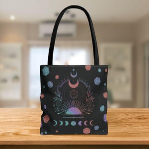 May include: A black tote bag with a pink and blue design featuring a 20-sided die, moons, and a sun. The bag is decorated with a repeating pattern of pink and blue geometric shapes.