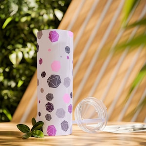 May include: A white tumbler with a geometric pattern of pink, purple, and gray shapes. The tumbler has a clear lid and straw. The design is repeated around the cylindrical body of the tumbler. The tumbler is on a wooden surface.
