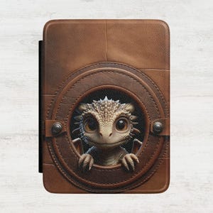 May include: A brown rectangular e-reader case featuring a circular porthole design. A small, textured, light-colored dragon with large dark eyes and spiky scales is peering through the center opening, resting its claws on the bottom edge.