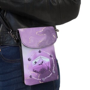 Könnte beinhalten: Violette Umhängetasche für Telefone mit Klappe. Die Tasche hat ein Fantasy-Design mit einer Drachensilhouette, geometrischen Formen und einer Waldszene. Goldakzente und ein schwarzer Riemen vervollständigen das Design.
