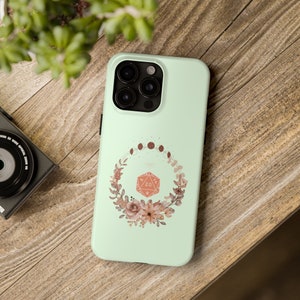 May include: A mint green phone case with a floral wreath design. The wreath surrounds a 20-sided die with a red and white design. The die is surrounded by a crescent moon and stars.