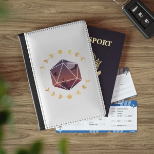 May include: A white passport holder with a black border and a gold design of a 20-sided die with a crescent moon pattern around it. The die has a sunset background with a silhouette of a castle and two dragons.