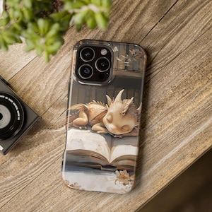 May include: A phone case with a cute illustration of a small, light brown dragon sleeping on an open book. The dragon has closed eyes and a light brown body with a light brown and white wing. The book is open and has a light brown cover with white pages. The phone case has a black camera lens with four lenses.