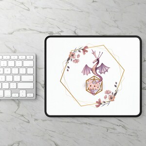 Könnte beinhalten: Eine weiße Mausmatte mit schwarzem Rand zeigt einen rosa und violetten Drachen mit goldenen Flügeln in einem goldenen geometrischen Rahmen. Der Drache ist von rosa Blumen umgeben.