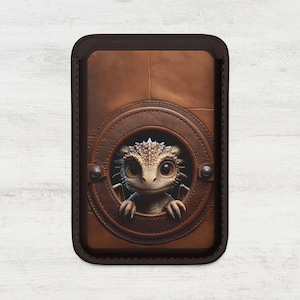 May include: A brown leather card holder with a circular window showcasing a cute dragon. The dragon has large eyes and a spiky head. The card holder has a dark brown border and is stitched.