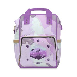 Puede incluir: Mochila morada con un diseño de acuarela que presenta un dado morado y dorado con un dragón volando sobre una cordillera. El dado está rodeado de dados más pequeños.