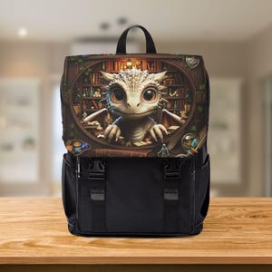 May include: Black backpack featuring a whimsical illustration of a baby dragon in a library setting. The dragon has large, expressive eyes and is surrounded by books and magical items. The backpack has a black base with adjustable straps and buckles.