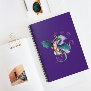 Peut inclure: Un carnet violet avec une illustration aquarelle d'un dragon vert et violet avec des accents dorés. Le dragon vole avec ses ailes déployées et est entouré de formes géométriques.