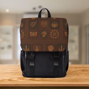 May include: A black backpack with a brown flap decorated with icons such as a sword, dice, and a shield. The flap has a stitched leather effect. The backpack has two black adjustable straps and two black buckles.