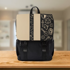Könnte beinhalten: Ein schwarzer Rucksack mit einer beigefarbenen Klappe, die einen vertikalen Streifen mit goldfarbenen Symbolen aufweist. Die Symbole umfassen ein Schwert, einen Schild, eine Laute, eine Faust und einen Würfel.