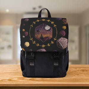 Può includere: Zaino nero con patta con un design geometrico di un dado D20, circondato da un motivo dorato a forma di luna crescente e stelle. Forme geometriche e lune aggiuntive sono sparse sullo sfondo nero. Lo zaino ha cinghie e fibbie nere.