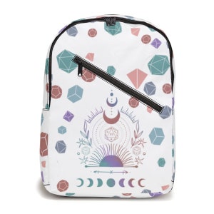 May include: White backpack with a colorful pattern of dice and celestial symbols. The design features a large 20-sided die, a crescent moon, a sun, and a row of moons.