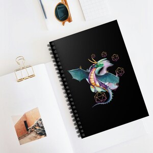 May include: A spiral notebook with a black cover featuring a colorful dragon with geometric shapes in pink and yellow. The dragon is flying with its wings spread out.
