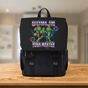 Puede incluir: Mochila negra con solapa que presenta un gráfico de tres figuras verdes en posturas de yoga, con el texto "GOBLIN YOGA MASTER" en degradado. La mochila tiene correas y hebillas ajustables.