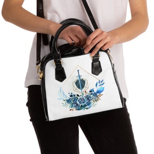Puede incluir: Un bolso de mano de cuero negro con un diseño blanco que presenta una forma geométrica azul y dorada con una espada en su interior, rodeada de flores azules.