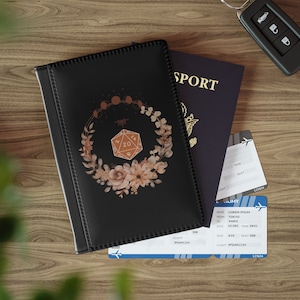 Könnte beinhalten: Ein schwarzer Reisepass-Etui mit einem floralen Design mit einem 20-seitigen Würfel und einem Halbmond. Das Etui liegt neben einem blauen Reisepass mit dem Wort "SPORT" auf dem Cover und zwei Flugtickets.