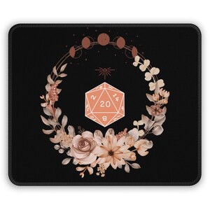Peut inclure: Un tapis de souris noir avec un motif de couronne de fleurs dans des tons de rose et de brun. La couronne entoure un dé à 20 faces avec les chiffres de 1 à 20. Le dé est de couleur pêche avec des chiffres blancs. Il y a six croissants de lune dans la couronne et une étoile au centre.