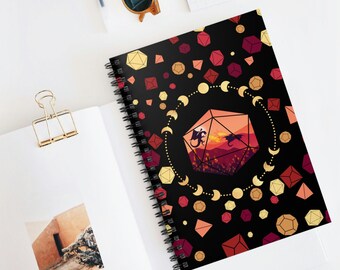 May include: A black spiral notebook with a colorful design featuring a 20-sided die with a sunset scene and two dragons flying over a mountain range. The die is surrounded by a circle of gold crescent moons and red, orange, and gold dice.