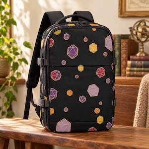 May include: A black backpack featuring an all-over print of multi-colored polyhedral dice in shades of purple, yellow, orange, and red. The bag includes adjustable side compression straps, a top handle, and padded shoulder straps.