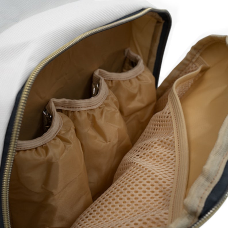 May include: A white nappy bag with a zipped opening, revealing a beige interior with multiple pockets and a mesh compartment. The bag has a black trim and gold zipper.