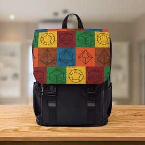 Può includere: Zaino nero con un colorato motivo geometrico di dadi sul risvolto. Il motivo presenta varie forme di dadi in una griglia di quadrati rossi, arancioni, gialli, verdi e blu. Lo zaino ha un manico e spallacci neri.