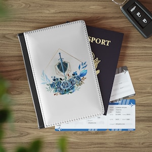 May include: A white passport holder with a black border and a blue floral design featuring a sword and shield. The holder is next to a blue passport with the word "SPORT" on the cover and a boarding pass with the text "LOREM IPSUM" and "IPSUM1234".