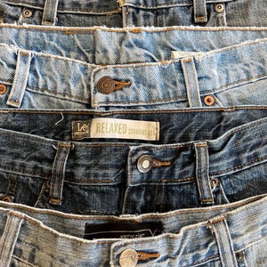May include: Close-up of several pairs of denim jeans in various shades of blue. The jeans have visible details like button closures, belt loops, and stitching. One pair has a "Lee Relaxed Straight Leg" label.