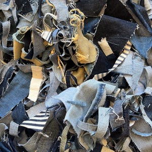 May include: A pile of denim fabric scraps in various shades of blue, including light, medium, and dark washes. The scraps are cut into irregular shapes and sizes, with some pieces showing frayed edges and visible stitching. The texture is rough and varied.