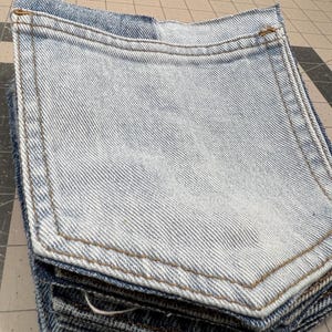 May include: A stack of light blue denim pockets with brown stitching. The pockets are rectangular with rounded corners and a curved top edge. The denim has a faded, worn appearance.