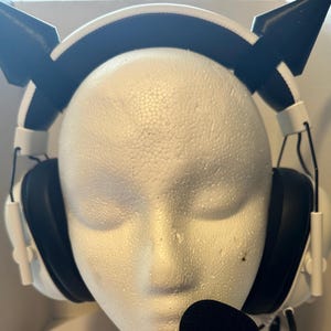 May include: White gaming headset with black earcups and a black microphone. The headset features black cat ear attachments on the headband. The headset is displayed on a white mannequin head.