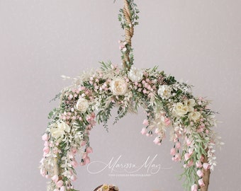 May include: A newborn baby girl sleeping in a white, fluffy nest, suspended from a floral wreath. The wreath is made of pink and white flowers and greenery, and is hanging from a rope attached to a wooden branch. The text "Marissa May Tiny Cuddlers Photography" is visible below the wreath.