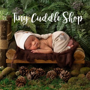 Puede incluir: Un bebé recién nacido con un gorro blanco y pantalones beige duerme en una cama de madera con una manta marrón. La cama está rodeada de vegetación verde y piñas.