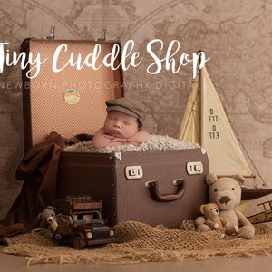May include: A newborn baby wearing a brown cap is lying in a brown suitcase with a brown blanket. The suitcase is surrounded by a brown fabric, a toy truck, a toy airplane, and two teddy bears. The background is a world map.