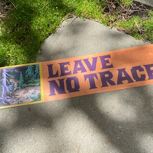 May include: A brown sign with the text "LEAVE NO TRACE" in purple letters. The sign has a small illustration of a forest scene with a path.