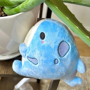 May include: A blue plush toy shaped like a whale with a black eye and a white belly. The whale is sitting on a wooden surface next to a potted aloe vera plant.