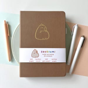 May include: A brown A5 notebook with a gold capybara illustration. The notebook has a white band with the text "soobiumi bullet journaling" and details about the pages. Two white pens and a peach pen are also visible.
