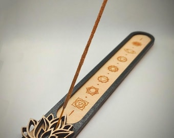 May include: A wooden incense holder with a lotus flower design and chakra symbols. The holder is black with a natural wood inlay. An incense stick is lit and placed in the holder.