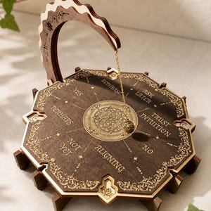 Könnte beinhalten: Ein Pendelbrett aus dunklem Holz mit einem bogenförmigen Ständer und einem hängenden Metallpendel. Das Brett zeigt ein zentrales Mandala-Design und Texte wie YES, PERHAPS, NOT YET, ALIGNING, NO, TRUST INTUITION, ASK LATER und REPHRASE.