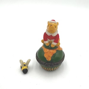 May include: A Winnie the Pooh figurine in a Santa suit sits atop a green and orange decorative box. A small bee figurine is next to the box. The box has the word "Winnie" on it.