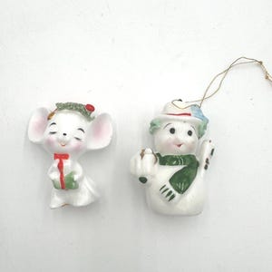 May include: Two ceramic Christmas ornaments. One is a white mouse with a red bow and a green gift. The other is a snowman with a green scarf and a hat. Both ornaments have gold string hangers.