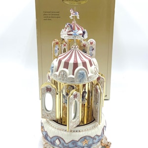 May include: A Gold Label Collection musical carousel featuring a red and white striped roof, gold accents, and miniature horses. The base is cream-colored with blue scrollwork and gold stars. The box text reads: Carousel turns and plays 16 Christmas carols as doors open and close.