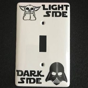 May include: White light switch cover with a black and white illustration of Baby Yoda on the top and the text "Light Side". On the bottom is a black and white illustration of Darth Vader and the text "Dark Side".