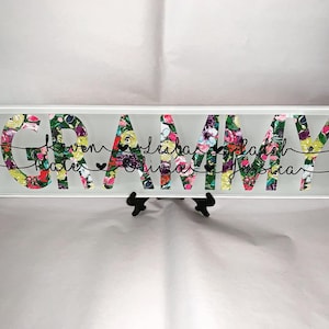 May include: A white rectangular sign with the word "GRAMMY" in black letters. The letters are filled with a floral pattern in shades of pink, green, yellow, and purple. The names "Karen, Katie, Olivia, Jessica, Leah, and Jadah" are written in black cursive script below the word "GRAMMY".