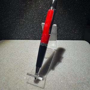 May include: A ballpoint pen featuring a bright red marbled barrel and a glossy black lower section. The pen includes polished chrome accents, a textured center band, and a silver pocket clip, displayed on a clear acrylic stand.