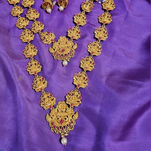 May include: A gold-tone necklace set with a double strand of pendant charms featuring a deity figure. The charms are adorned with red gemstones and the necklace has a pearl drop. The earrings are matching.